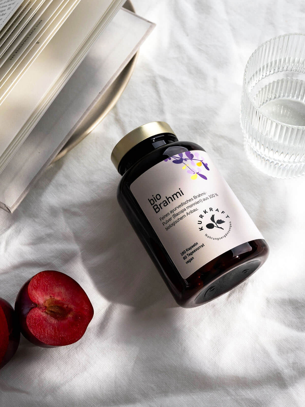 Kurkraft Bio Brahmi Kapseln in Dose auf weißem Tuch mit Pflaumenhälften, Buch und Wasserglas