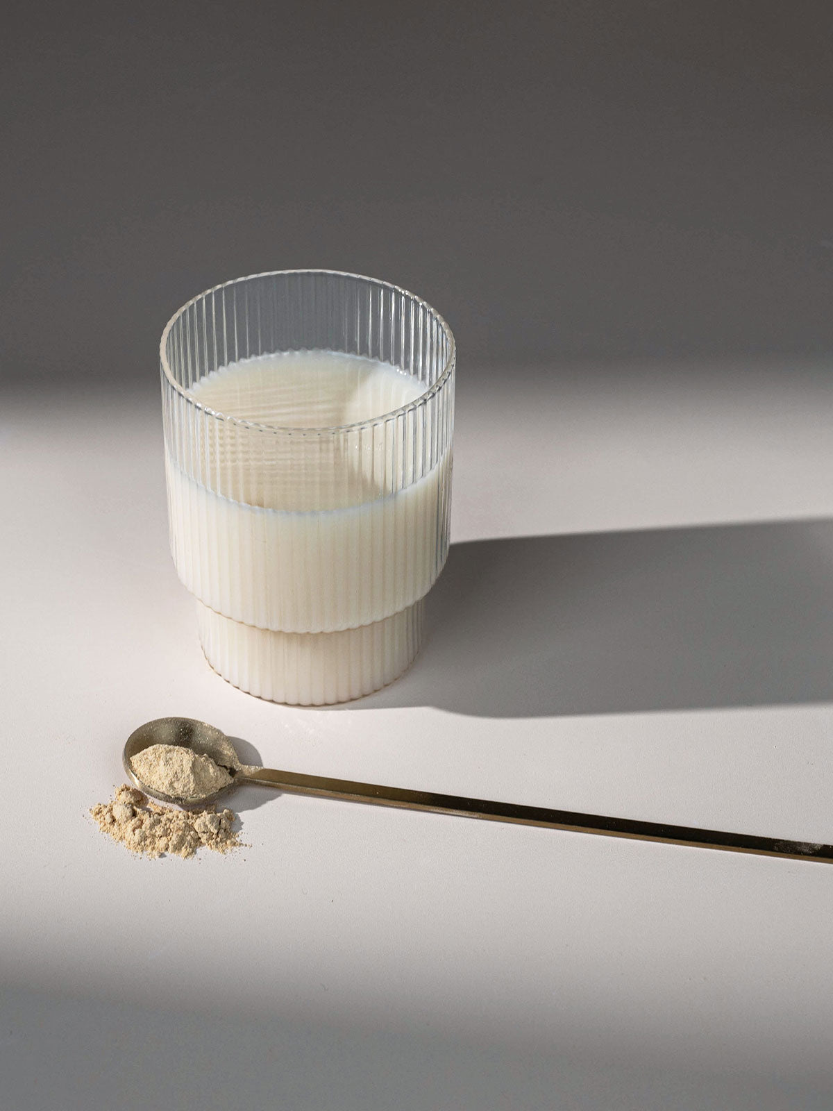 Glas mit Ashwagandha-Pulver in Milch angerührt auf hellem Untergrund mit goldenem Löffel