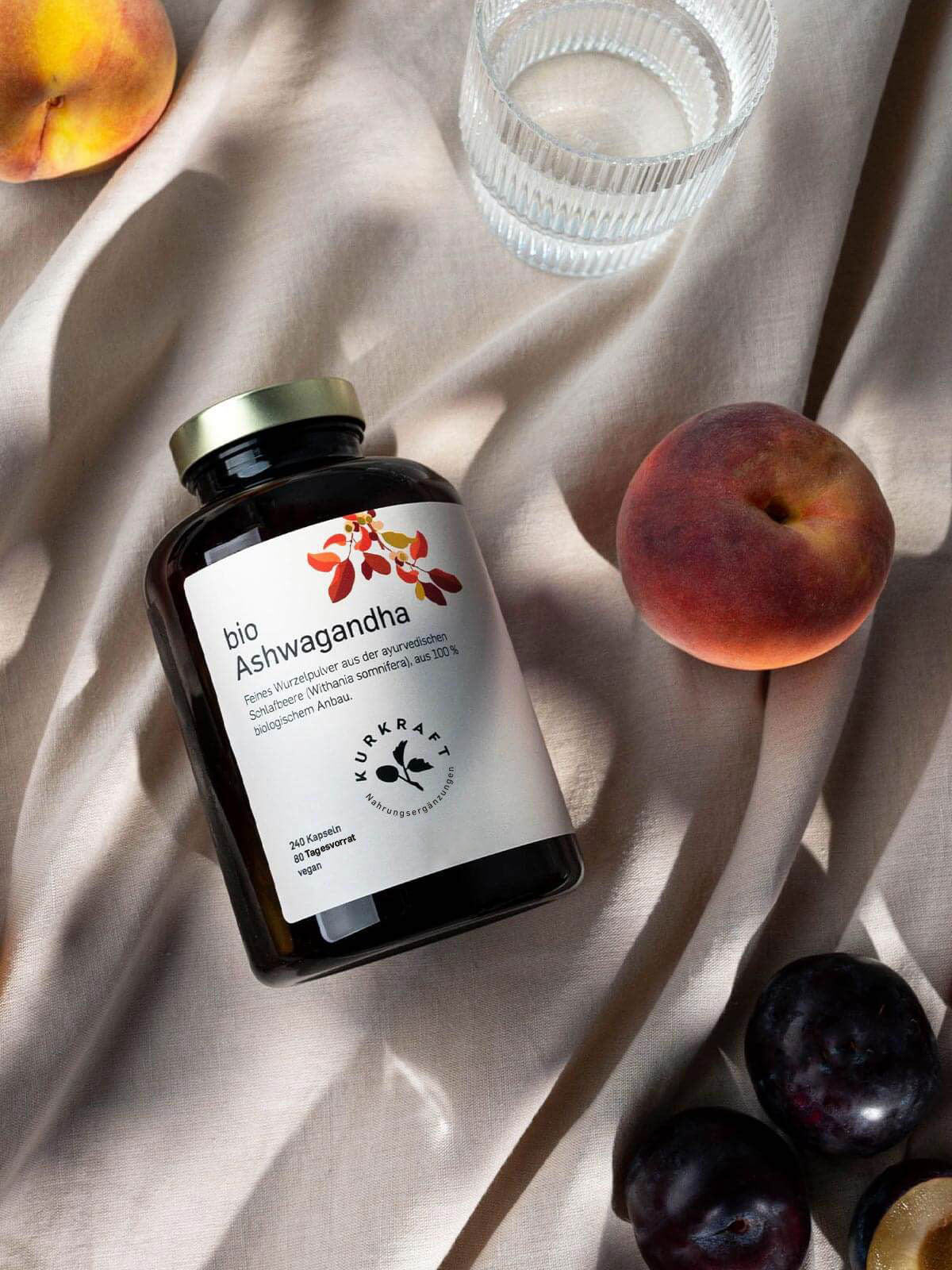 Kurkraft Bio Ashwagandha Kapseln mit Glas Wasser auf hellem Stoff – hochwertiges Adaptogen aus der ayurvedischen Wurzel Withania somnifera.