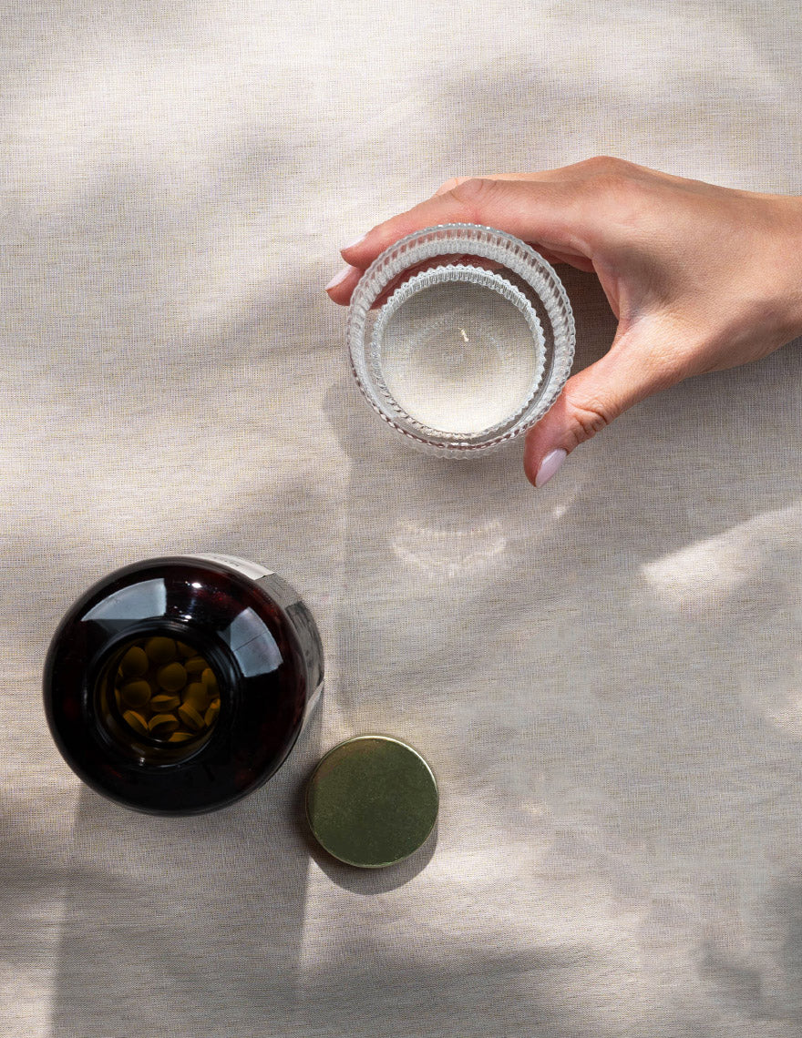 Hand hält ein Glas Wasser neben einer geöffneten Flasche mit Bio Ashwagandha Tabletten von Kurkraft auf hellem Leinentuch.