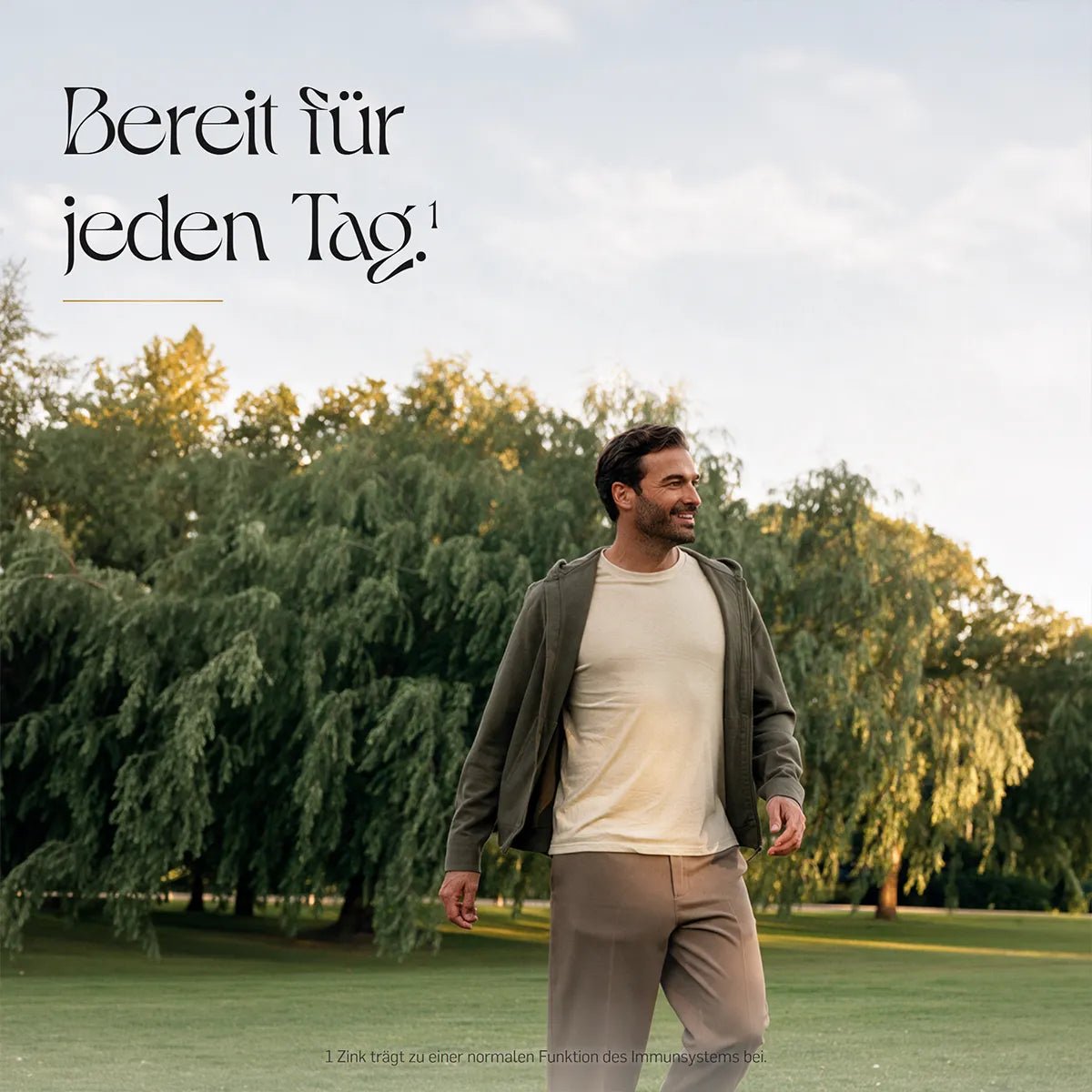 Bereit für jeden Tag – lächelnder Mann in hellem Shirt und olivgrüner Jacke geht über eine Rasenfläche vor Trauerweiden im warmen Abendlicht.